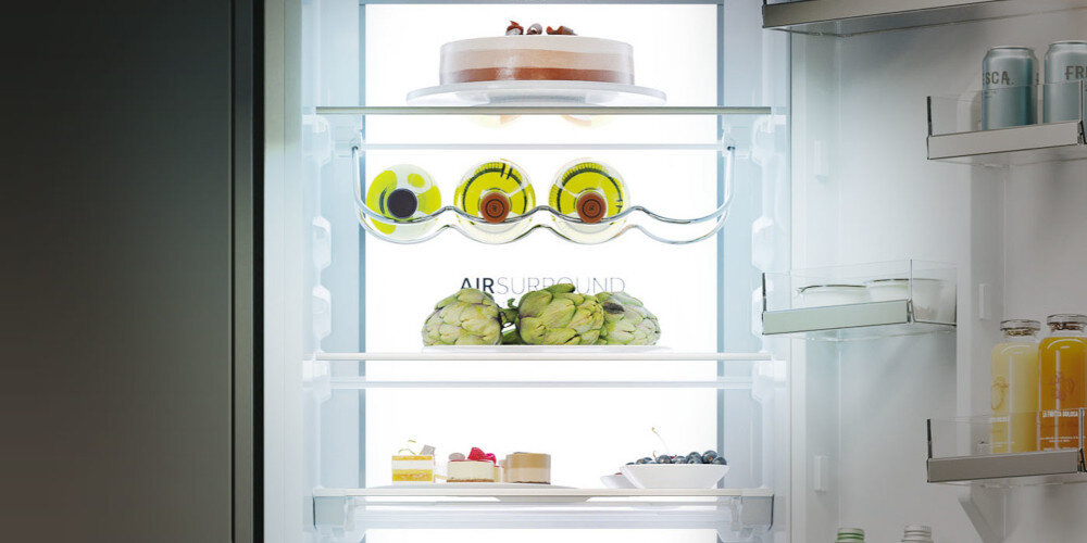 Wnętrze lodówki z oświetleniem, na półkach znajdują się ciasto, owoce, karczochy i napoje. Na drzwiach widoczne są dodatkowe półki z butelkami i słoikami.