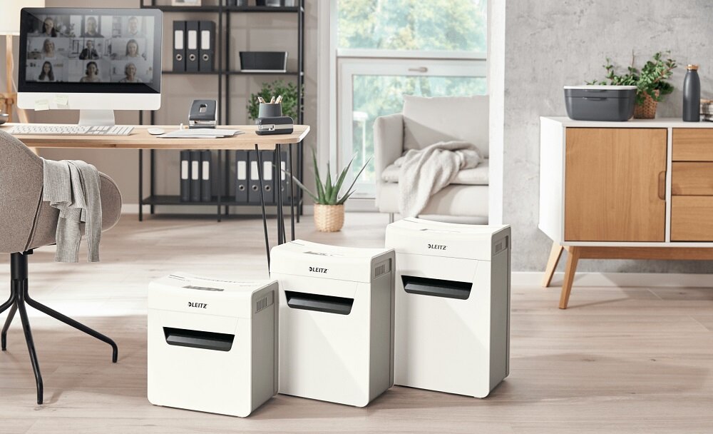 Trzy białe niszczarki z logo \'LEITZ\' stoją na podłodze w nowoczesnym biurze. W tle widać biurko z komputerem, półki z segregatorami i okno z widokiem na zieleń.