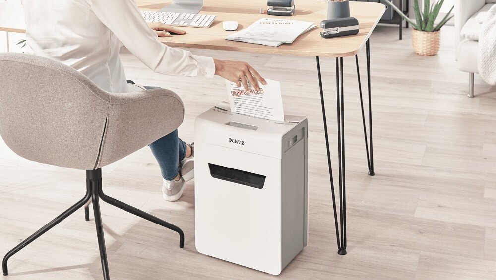 Osoba wkłada dokument do białej niszczarki z logo LEITZ, stojącej obok biurka. Na biurku znajdują się różne przedmioty biurowe, a w tle widać roślinę doniczkową.