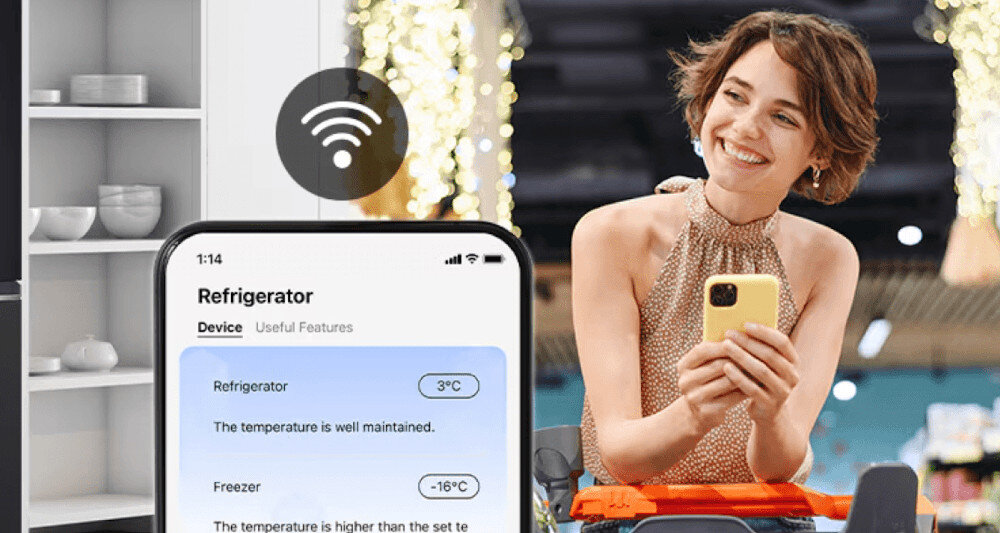 Na obrazie widać kobietę trzymającą telefon z aplikacją pokazującą temperatury: \'Refrigerator 3°C\' i \'Freezer -16°C\'. W tle znajduje się kuchnia z otwartą lodówką i ikoną sygnału Wi-Fi.