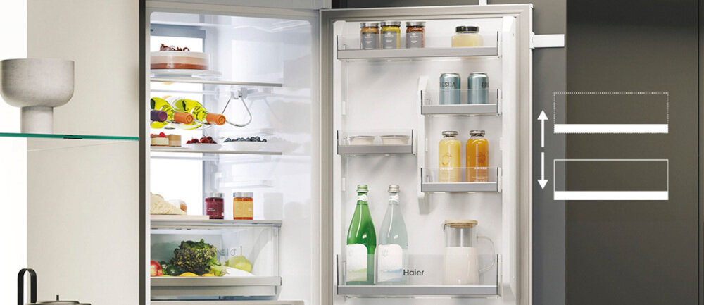Otwarta lodówka z widocznymi półkami wypełnionymi różnymi produktami spożywczymi, takimi jak butelki, słoiki i warzywa. Na drzwiach lodówki widoczne jest logo \'Haier\'.