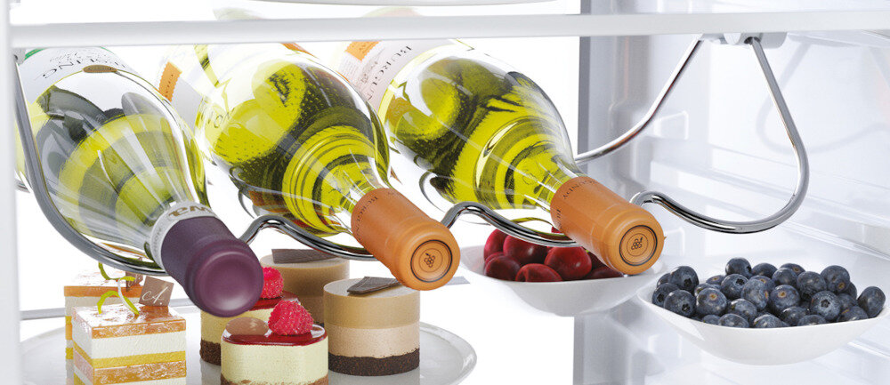 W lodówce znajdują się trzy butelki wina na metalowym stojaku oraz talerze z deserami, wiśniami i jagodami. Wnętrze jest jasno oświetlone.