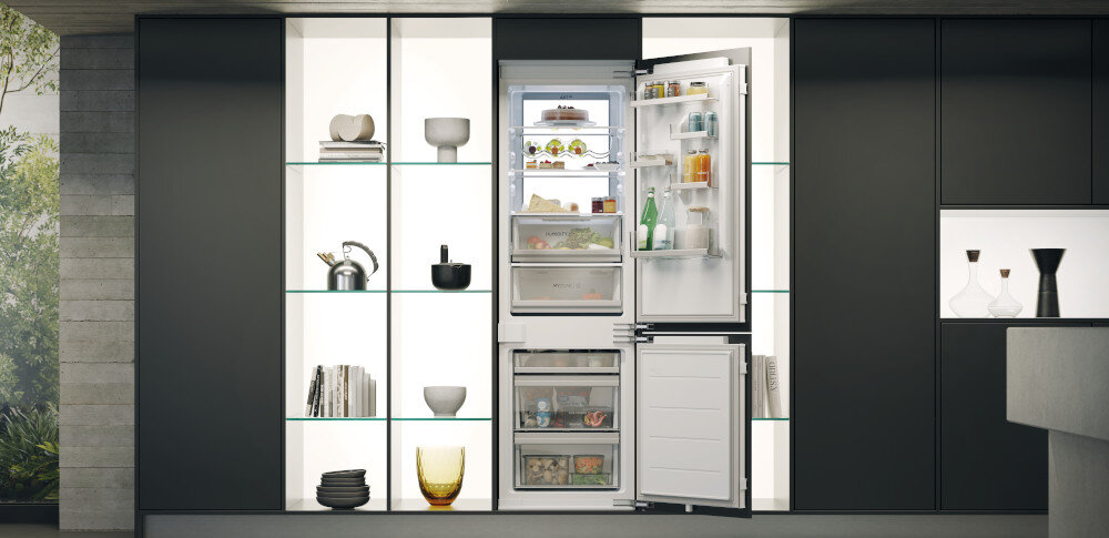 Wnętrze nowoczesnej kuchni z otwartą lodówką w zabudowie, wypełnioną różnorodnymi produktami spożywczymi. Obok lodówki znajdują się półki z dekoracjami i książkami.