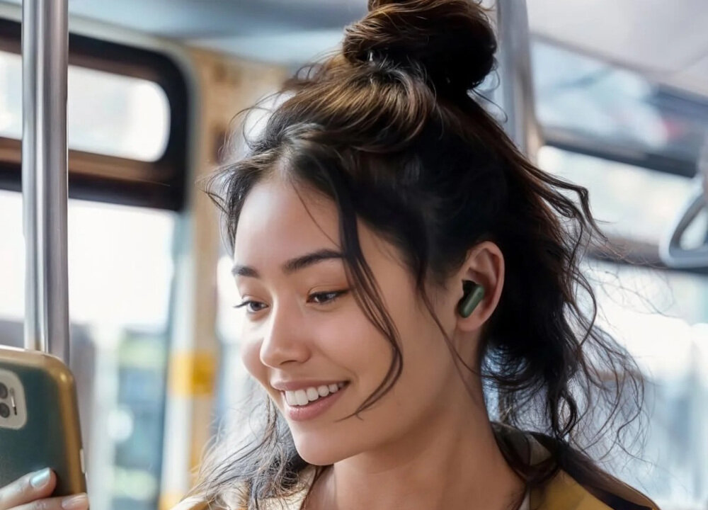 Osoba nosi zielone słuchawki dokanałowe. W tle widać wnętrze pojazdu komunikacji miejskiej.