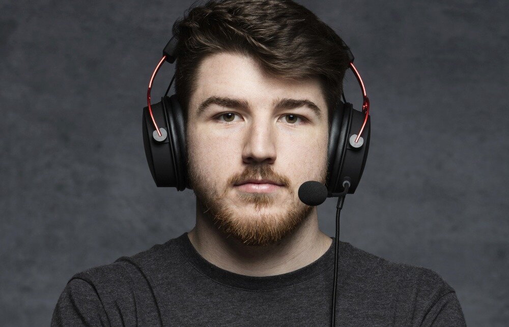 Osoba nosi czarne słuchawki nauszne z czerwonymi elementami i mikrofonem. Tło jest szare i jednolite.