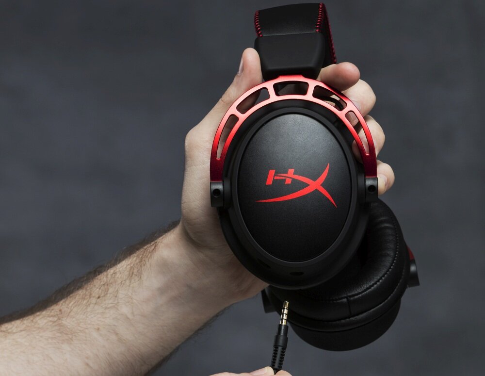 Osoba trzyma czarne słuchawki z czerwonymi elementami i logo \'HX\' na nauszniku. Widać również wtyczkę jack zbliżającą się do słuchawek.