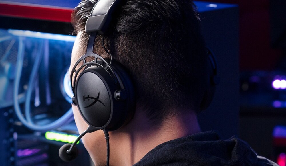 Osoba nosi czarne słuchawki nauszne z widocznym logo \'HyperX\'. W tle znajduje się komputer z podświetleniem LED.