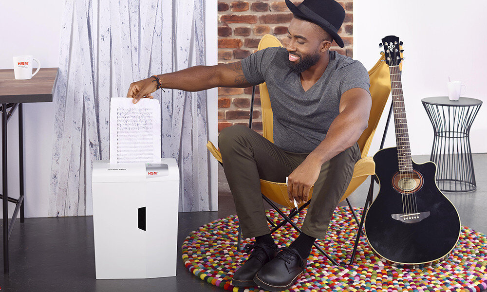 Osoba wkłada kartkę z tekstem do niszczarki HSM Shredstar. Obok stoi gitara akustyczna, a w tle widać ceglaną ścianę i zasłonę.