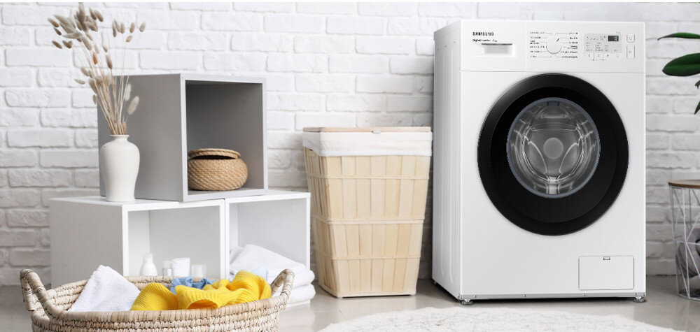 Na obrazie znajduje się pralka z widocznym logo \'SAMSUNG\', obok której stoi kosz na pranie oraz półka z dekoracjami. W tle widoczna jest biała ceglana ściana.