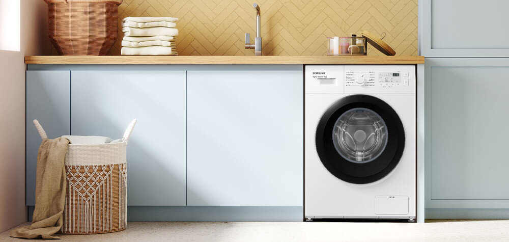W kuchni znajduje się pralka w zabudowie z widocznym panelem sterowania i logo SAMSUNG. Obok pralki stoi kosz na pranie, a na blacie leżą złożone ręczniki.