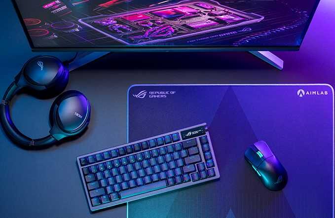 Na biurku znajduje się klawiatura mechaniczna, mysz komputerowa oraz słuchawki, obok monitora wyświetlającego grafikę technologiczną. Na podkładce pod mysz widoczne są logotypy \'Republic of Gamers\' i \'AIMLAB\'.