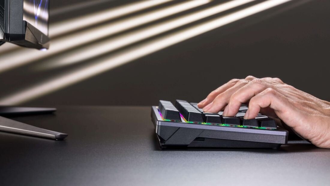 Osoba pisze na czarnej klawiaturze mechanicznej z podświetleniem RGB. W tle widoczny jest fragment monitora na ciemnym biurku.