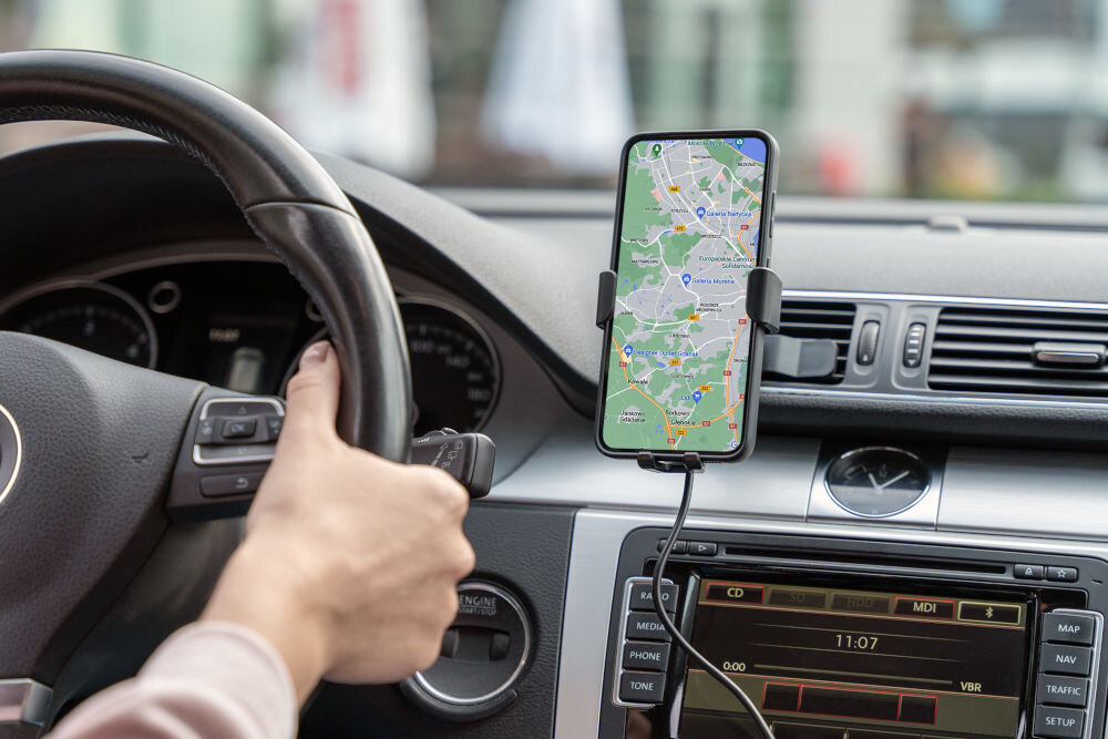 Wnętrze samochodu z zamontowanym uchwytem na telefon, w którym znajduje się smartfon z włączoną aplikacją mapową. Na ekranie radia widoczna jest godzina 11:07.