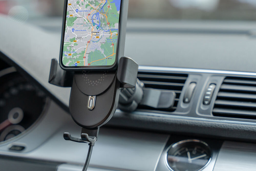 Czarny uchwyt na telefon zamontowany w samochodzie, trzymający smartfon z wyświetloną mapą. Uchwyt jest przymocowany do kratki wentylacyjnej deski rozdzielczej.