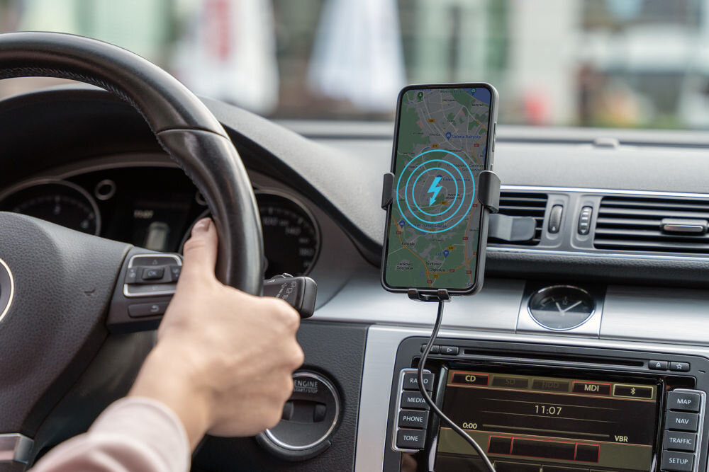 Wnętrze samochodu z zamontowanym uchwytem na telefon, w którym znajduje się smartfon z wyświetloną mapą i ikoną ładowania. Kierowca trzyma kierownicę, a na ekranie radia widoczna jest godzina 11:07.