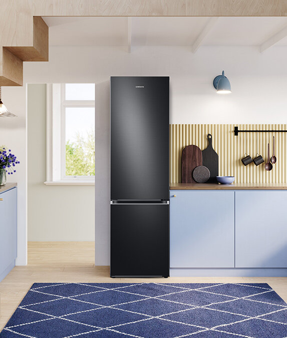 W kuchni znajduje się wysoka, grafitowa lodówka z logo Samsung. Obok niej są niebieskie szafki, a na blacie stoją deski do krojenia i miska.
