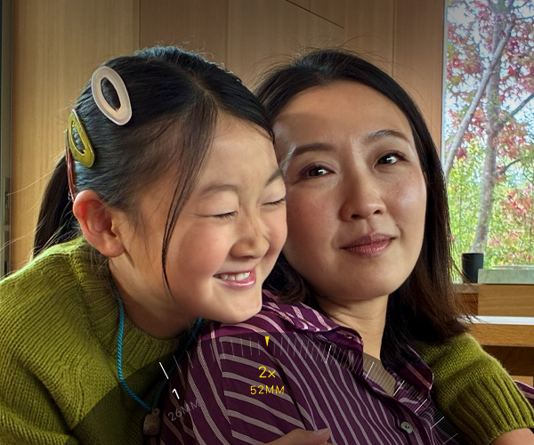 Na zdjęciu widać dwie osoby w pomieszczeniu, jedna z nich się uśmiecha. Na obrazie widoczny jest interfejs aparatu z oznaczeniem \'2x 52MM\'.