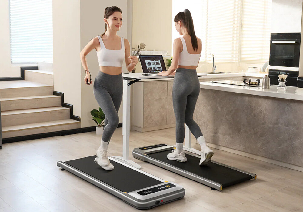 Dwie kobiety ćwiczą na bieżniach elektrycznych w nowoczesnej kuchni. Jedna z nich korzysta z laptopa umieszczonego na stojaku przed bieżnią.