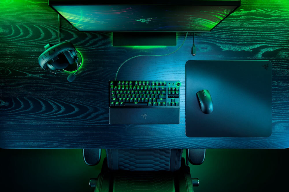 Na ciemnym biurku znajduje się klawiatura z podświetleniem, mysz oraz podkładka z logo Razer. Obok monitora wisi słuchawka na uchwycie, a całość oświetlona jest zielonym światłem.