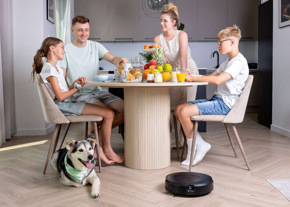 Wnętrze kuchni z okrągłym stołem, na którym znajdują się owoce, soki i pieczywo. Na podłodze widoczny jest robot sprzątający.