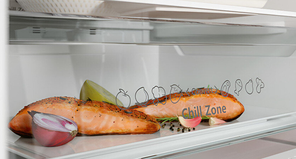 Wnętrze lodówki z półką, na której leży kawałek łososia, cebula i przyprawy. Na szufladzie widoczny jest napis \'Chill Zone\' oraz grafiki owoców i warzyw.