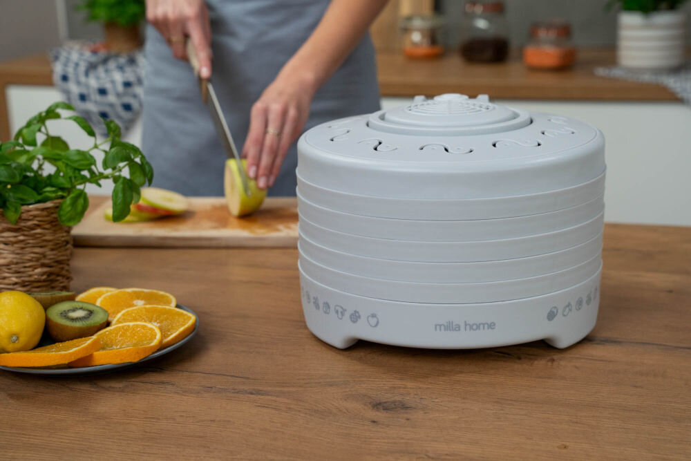 Suszarka do grzybów MILLA HOME FD101E Szara suszarka do grzybów i owoców Milla Home FD101E stoi na drewnianym blacie. Na talerzu obok leżą plasterki kiwi, pomarańcza i cytryna. W tle, ręce kroją jabłko, przygotowując je do suszenia.