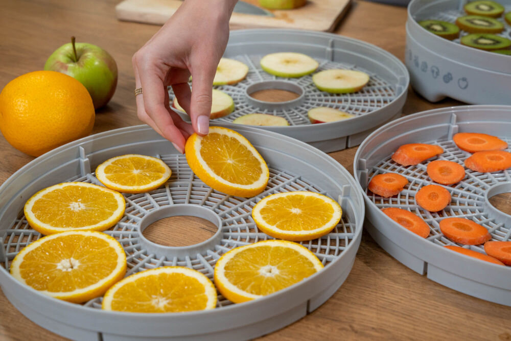 Suszarka do grzybów MILLA HOME FD101E Ręka układa plasterek pomarańczy na jednym z sit suszarki do żywności. Obok na innych sitach widoczne są plastry jabłek, kiwi oraz plasterki marchewki, leżące na drewnianym blacie.