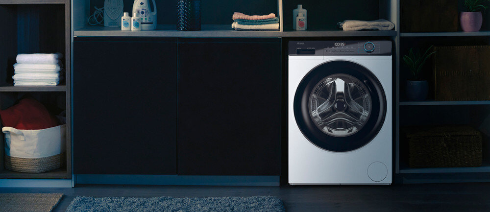 Pralka o nowoczesnym designie z dużym, okrągłym oknem bębna, umieszczona w eleganckiej łazience z ciemnymi szafkami. Na blacie znajdują się ręczniki i butelki z detergentami.