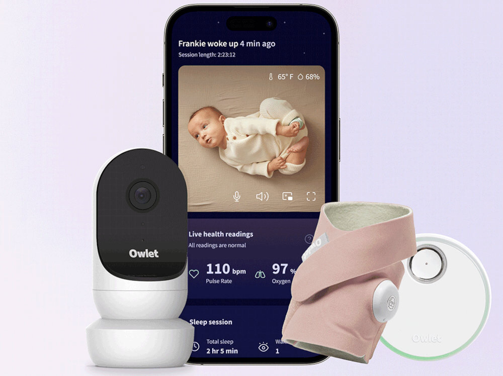 Na obrazie widoczna jest elektroniczna niania z kamerą Owlet oraz monitor oddechu w kolorze różowym. W tle znajduje się smartfon z aplikacją pokazującą obraz dziecka i dane zdrowotne, takie jak puls i poziom tlenu.