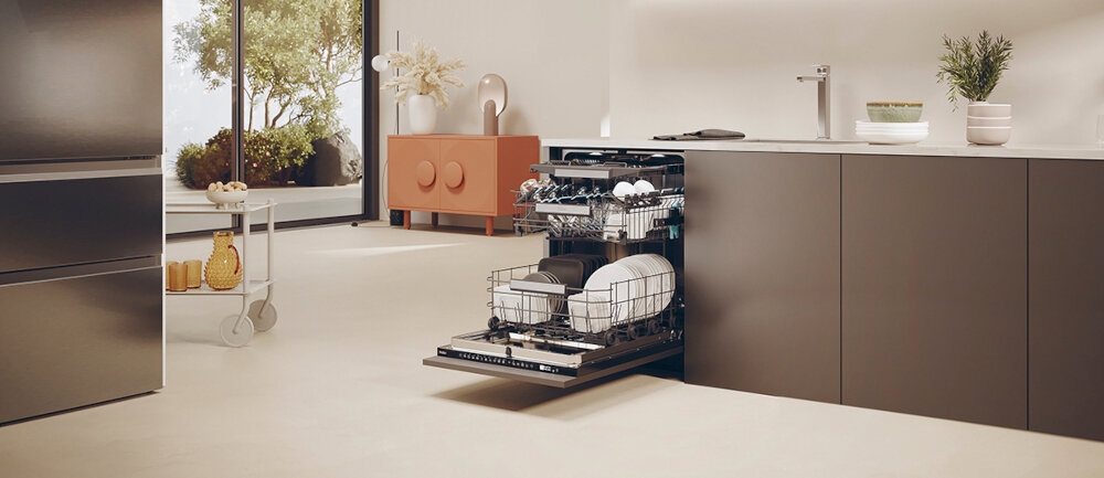W nowoczesnej kuchni znajduje się otwarta zmywarka z widocznymi naczyniami w środku. W tle widać szafkę z dekoracjami oraz wózek z naczyniami i rośliną.