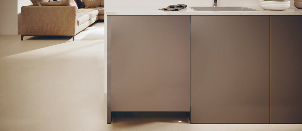 Nowoczesna kuchnia z zabudowaną zmywarką o gładkich, ciemnych frontach. W tle widoczna jest część salonu z jasną sofą.