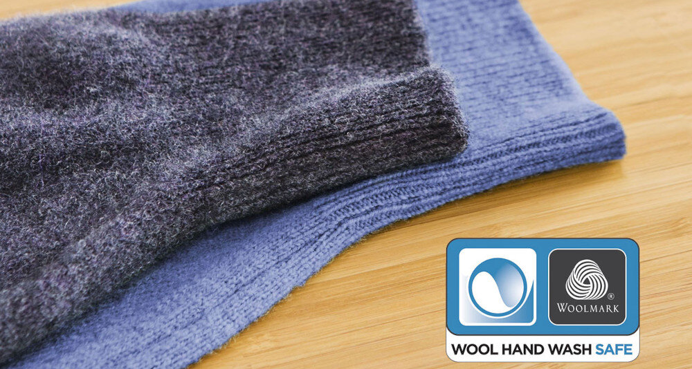 Na drewnianej powierzchni leżą dwa złożone swetry, jeden ciemnoszary, drugi niebieski. W prawym dolnym rogu znajduje się logo Woolmark z napisem \'WOOL HAND WASH SAFE\'.