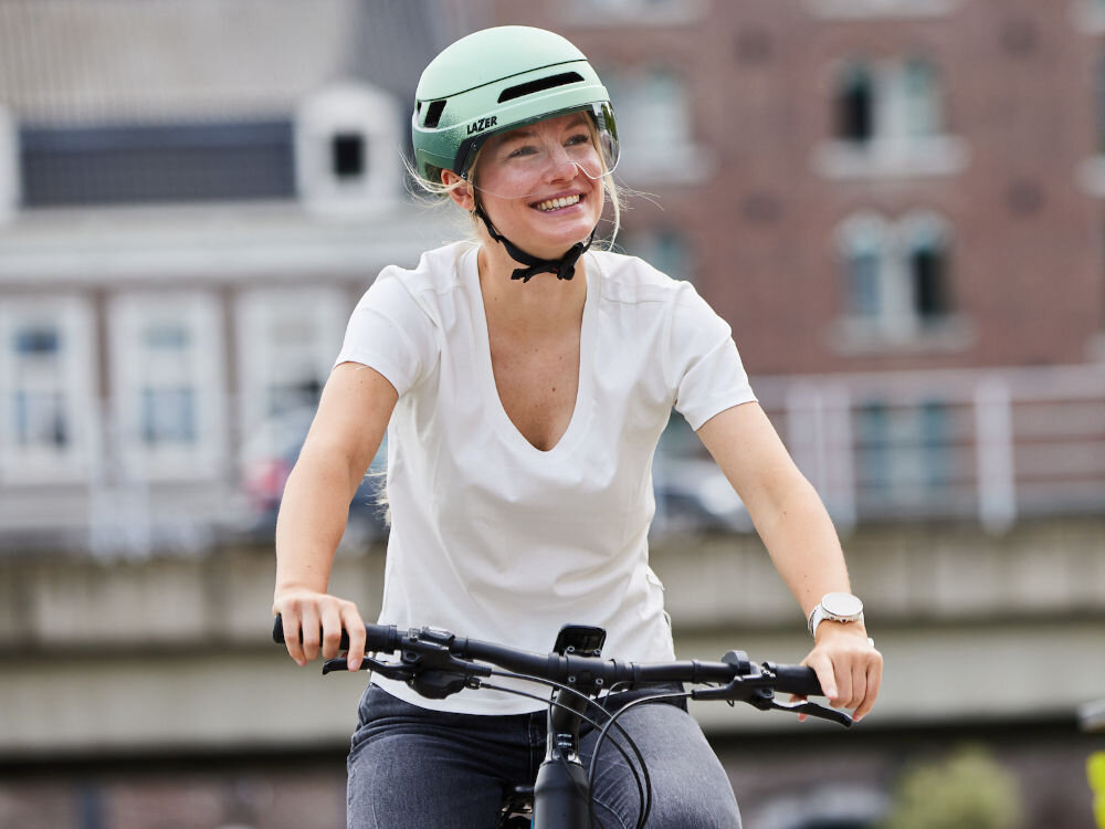 Osoba na rowerze nosi zielony kask z napisem \'LAZER\'. W tle widoczne są budynki i most.