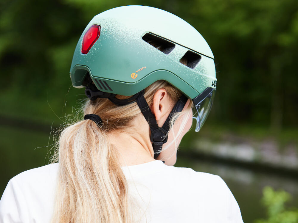 Osoba nosi zielony kask rowerowy z lampką z tyłu. Na kasku widoczne jest logo \'LAZER\'.