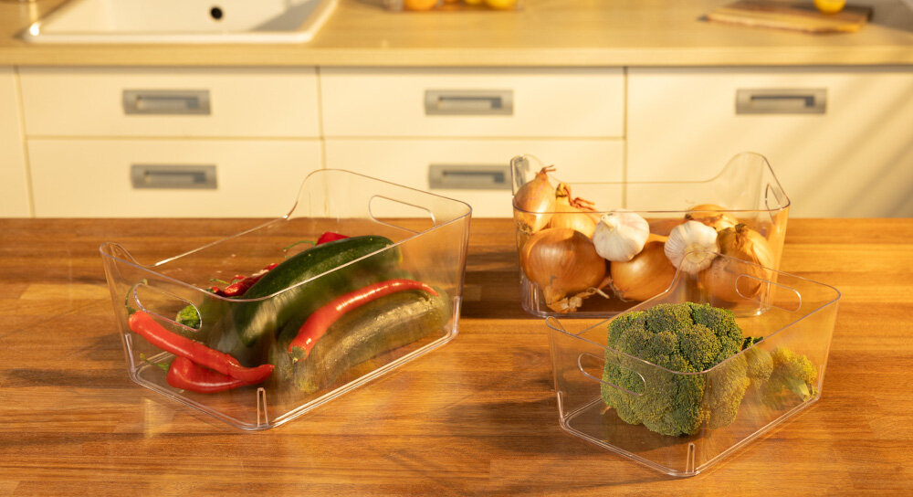 Na drewnianym blacie znajdują się trzy przezroczyste pojemniki z warzywami: jeden z paprykami i ogórkami, drugi z cebulą i czosnkiem, a trzeci z brokułem. W tle widoczna jest kuchnia z jasnymi szafkami i zlewem.