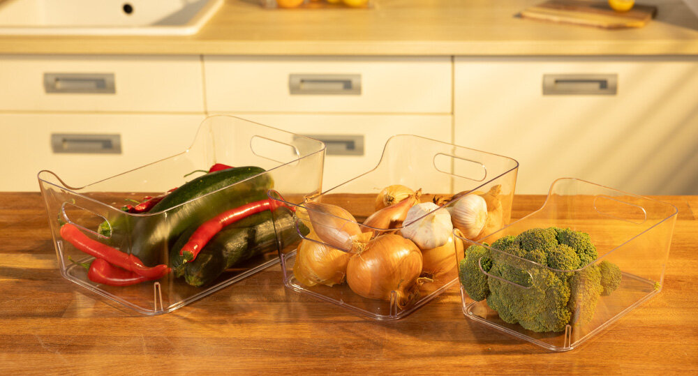 Na drewnianym blacie znajdują się trzy przezroczyste organizery wypełnione warzywami: papryką, ogórkami, cebulą, czosnkiem i brokułami. W tle widoczna jest kuchnia z jasnymi szafkami i zlewem.