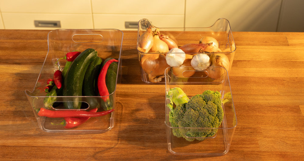 Na drewnianym blacie znajdują się trzy przezroczyste pojemniki z warzywami: papryki i ogórki, cebule i czosnek, oraz brokuł. Pojemniki mają wycięcia na uchwyty.