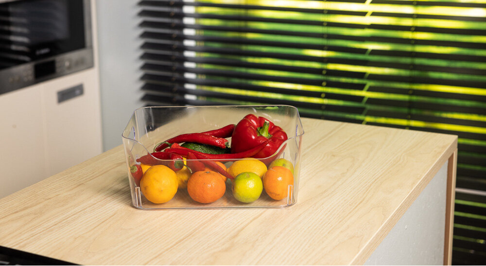 Na drewnianym blacie stoi przezroczysty pojemnik z owocami i warzywami, w tym papryką i cytrusami. W tle widoczne są żaluzje na oknie oraz fragment kuchni.