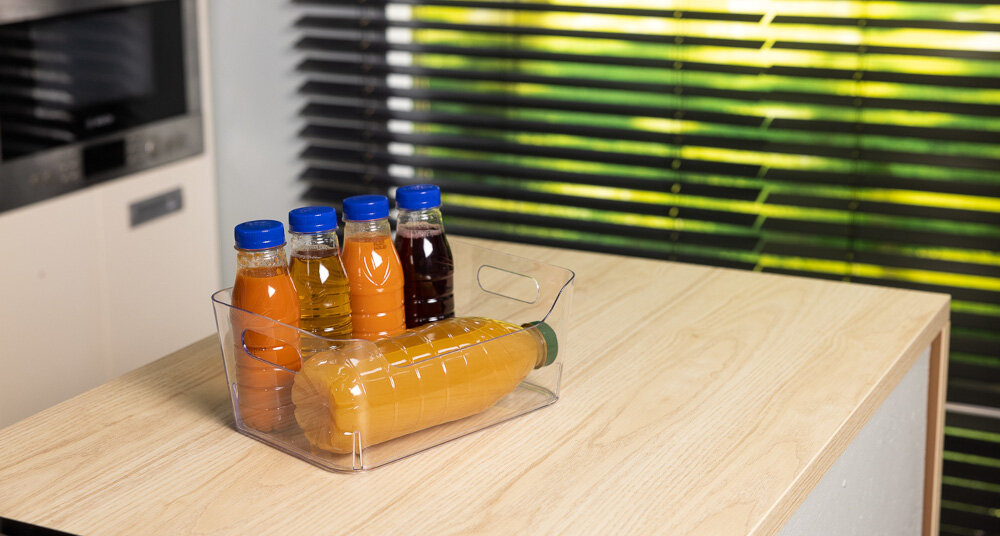 Na drewnianym blacie stoi przezroczysty organizer z kilkoma butelkami napojów o różnych kolorach i niebieskich zakrętkach. W tle widoczne są żaluzje na oknie.