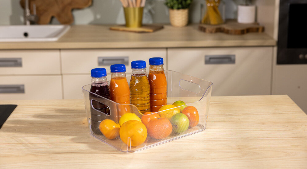 Na drewnianym blacie stoi przezroczysty organizer z trzema butelkami soku i kilkoma owocami, w tym pomarańczami i jabłkami. W tle widoczna jest kuchnia z blatem i zlewem.