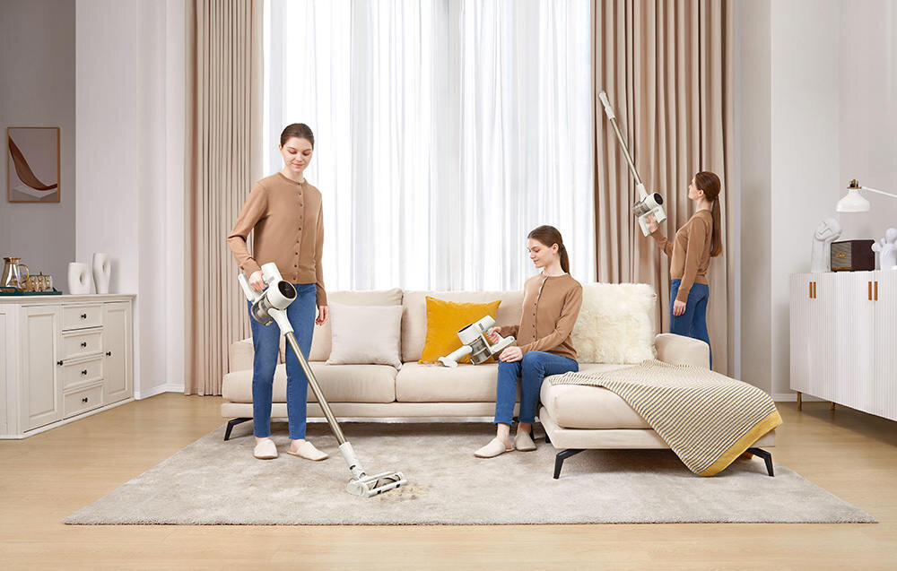 Trzy kobiety w pomieszczeniu używają odkurzacza pionowego na dywanie i przy zasłonach. W tle znajduje się sofa z poduszkami oraz szafka z dekoracjami.