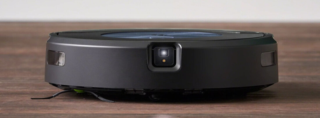 Na drewnianej podłodze znajduje się czarny robot sprzątający o okrągłym kształcie z widocznym czujnikiem z przodu. Robot ma gładką powierzchnię i jest w kolorze ciemnym.