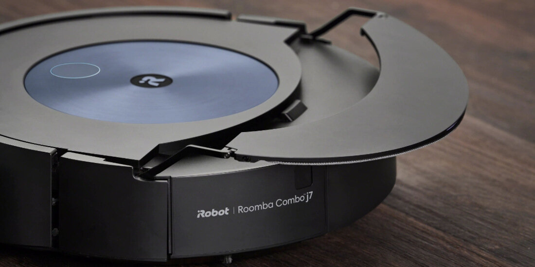 Na drewnianej powierzchni znajduje się robot sprzątający z widocznym logo i napisem \'iRobot Roomba Combo j7\'. Urządzenie ma okrągły kształt i ciemną kolorystykę.