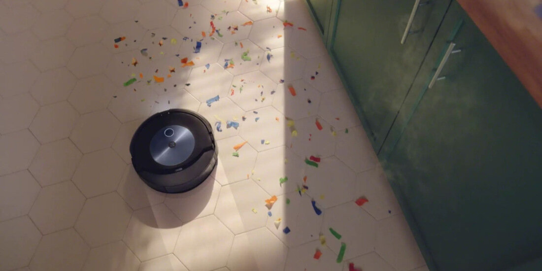 Na podłodze z jasnych płytek znajduje się robot sprzątający, otoczony kolorowymi kawałkami papieru. Obok widoczna jest szafka kuchenna z zielonymi frontami.