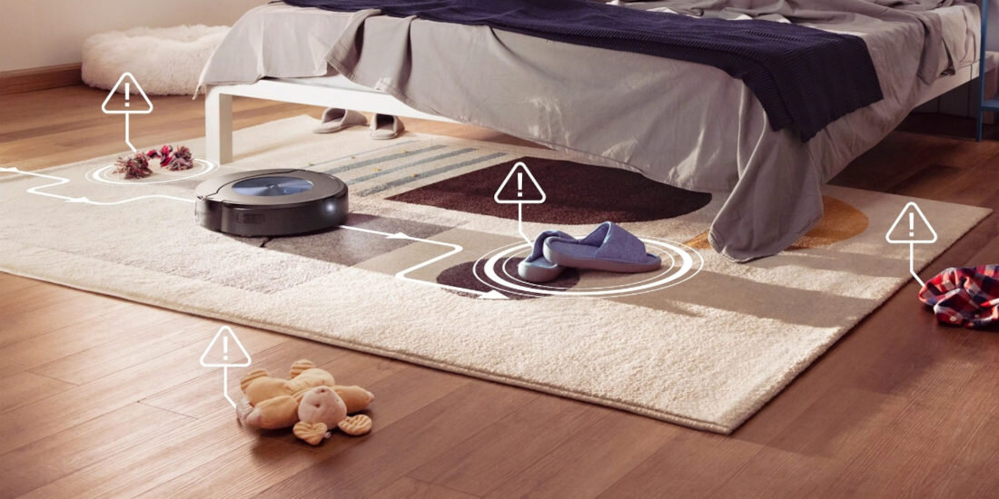 Robot sprzątający porusza się po dywanie w sypialni, omijając przeszkody takie jak zabawki i ubrania. Na dywanie widoczne są ikony ostrzegawcze wskazujące na przeszkody.