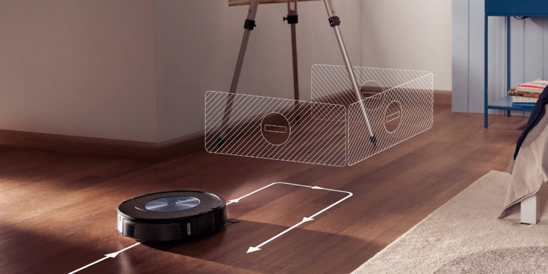 Robot sprzątający porusza się po drewnianej podłodze, a na jego trasie widoczne są białe strzałki wskazujące kierunek ruchu. W tle znajduje się przezroczysta bariera z oznaczeniami, sugerująca obszar, którego robot unika.