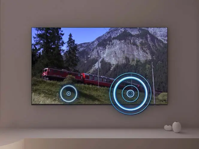 Na ścianie wisi telewizor wyświetlający obraz pociągu jadącego przez górski krajobraz. Na ekranie widoczne są dwa niebieskie okręgi symbolizujące dźwięk przestrzenny.