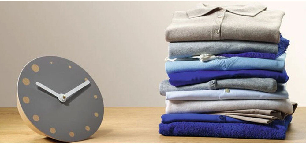 Na drewnianym blacie stoi okrągły zegar z szarym cyferblatem i złotymi kropkami zamiast cyfr. Obok znajduje się stos złożonych ubrań w różnych odcieniach niebieskiego i szarości.