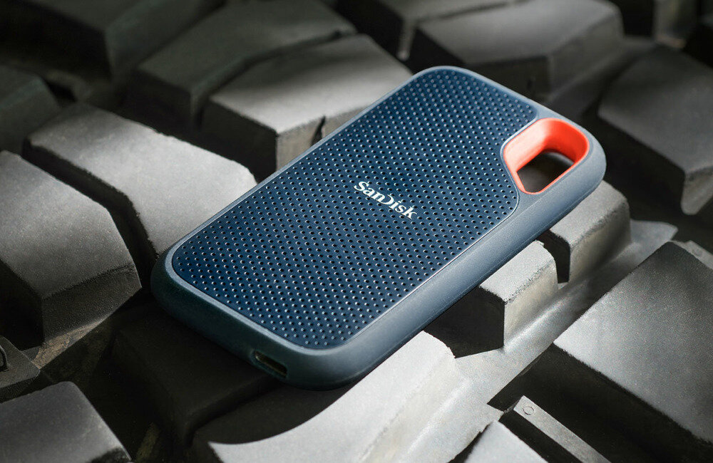 Na zdjęciu widoczny jest przenośny dysk SSD marki SanDisk z napisem \'SanDisk\' na obudowie. Dysk ma ciemnoniebieską powierzchnię z pomarańczowym akcentem przy otworze na smycz.