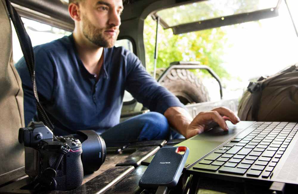 Mężczyzna pracuje na laptopie w pojeździe terenowym, obok znajduje się aparat fotograficzny i dysk SANDISK Extreme Portable SSD. Na dysku widoczne jest logo \'SanDisk\'.